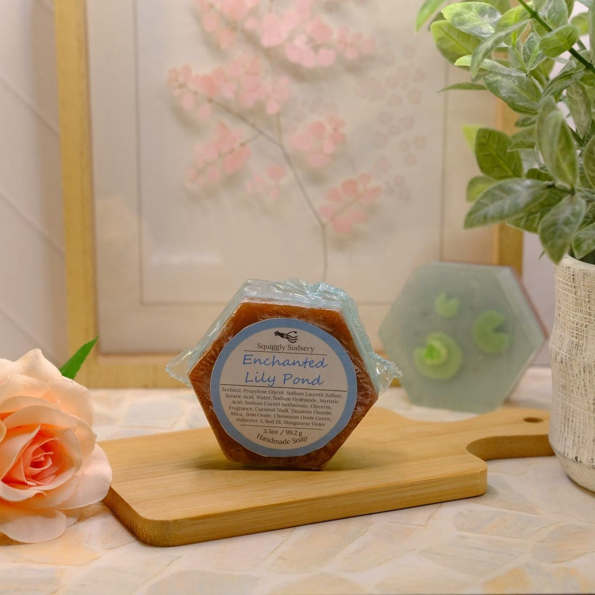 A hexagonal wrapped soap bar labeled 'Enchanted Lily Pond' sits on a wooden cutting board on a patterned tablecloth, styled with a peach rose, a potted plant, and a blurred soap bar in the background.