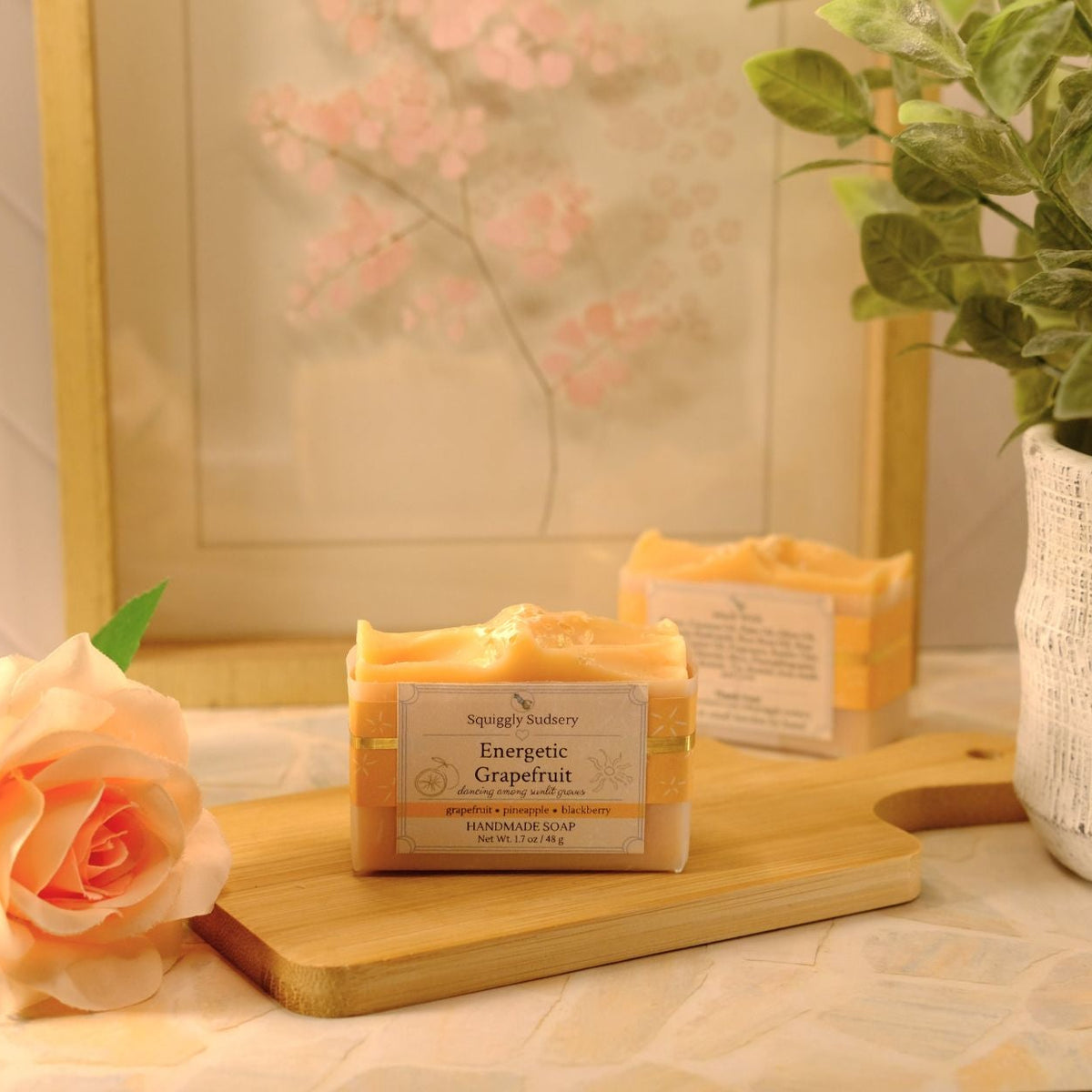 A bar of soap labeled 'Energetic Grapefruit' by Squiggly Sudsery sits on a wooden cutting board, styled with a peach rose, a potted green plant, and a framed print in a warm, cozy setting.
