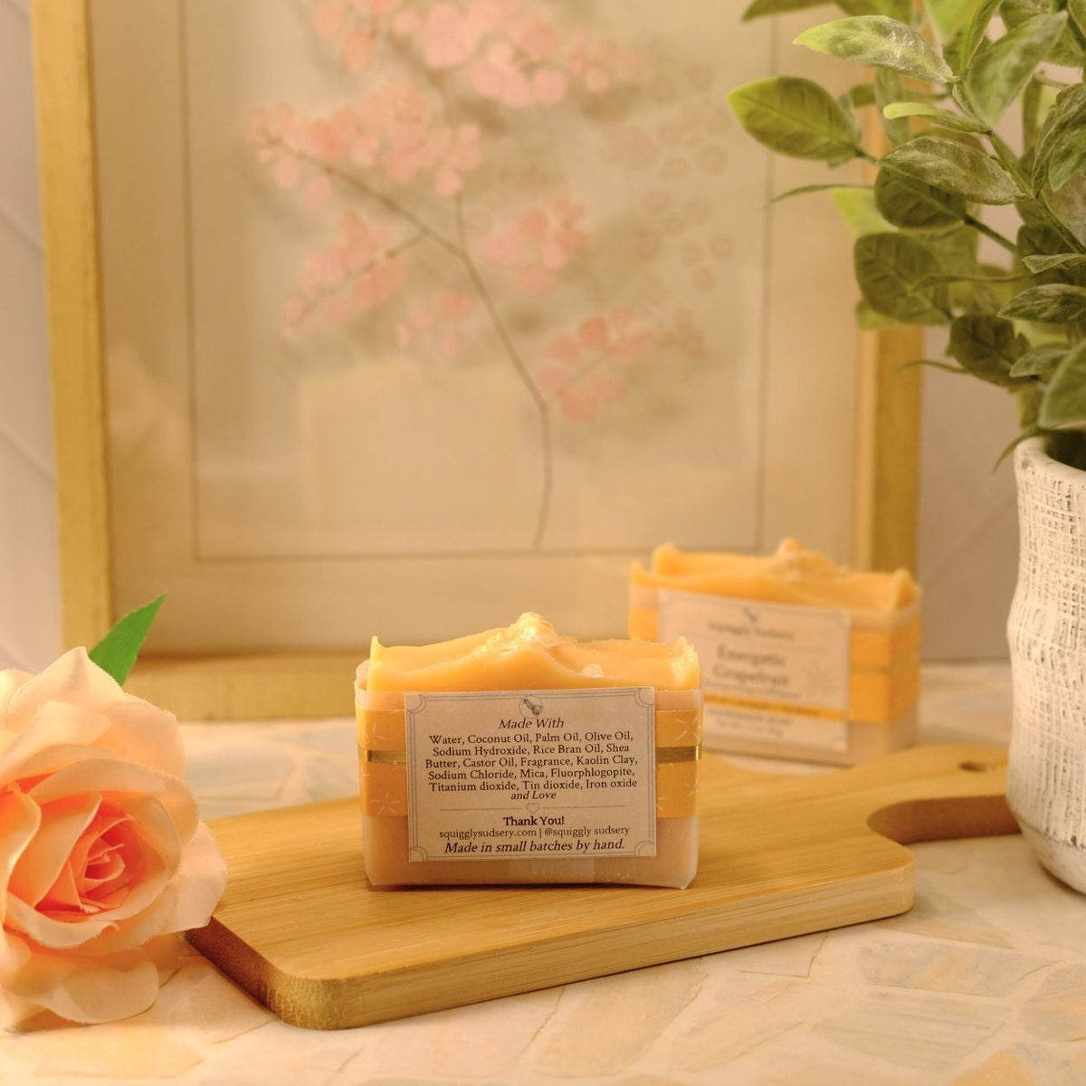 A bar of handmade soap sits on a wooden cutting board next to a peach rose, with another soap bar and a potted plant in the background.