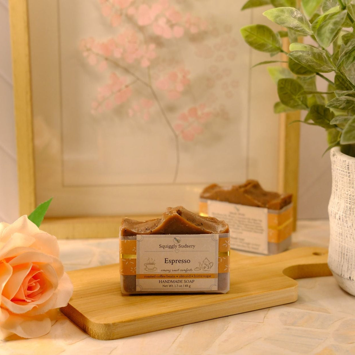 A bar of espresso scented soap sits on a wooden cutting board on a tabletop, styled with a peach rose, a potted green plant, and a framed floral print in the background.