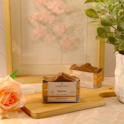 A bar of espresso scented soap sits on a wooden cutting board on a tabletop, styled with a peach rose, a potted green plant, and a framed floral print in the background.