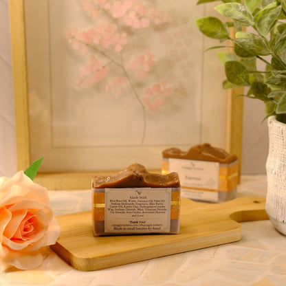 A rectangular bar of handmade soap with a brown and gold marbled top and a cream label, displayed on a wooden cutting board next to a peach rose, with another soap bar and a potted plant in the background.