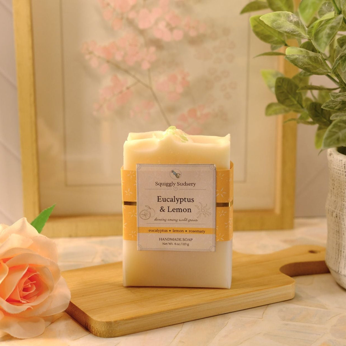 A rectangular bar of soap labeled 'Eucalyptus & Lemon' by Squiggly Sudsery, wrapped in a kraft paper band, displayed on a wooden cutting board with a peach rose and a potted green plant in a styled setting.