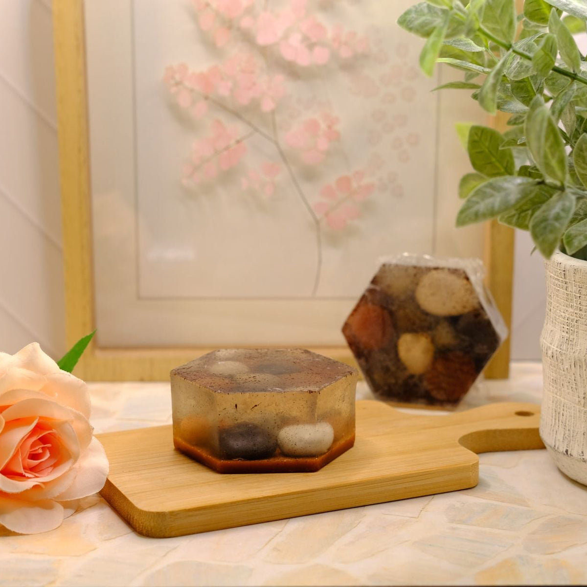Two hexagonal soaps with embedded stone-shaped soap, one resting on a wooden cutting board and the other in the background, styled on a table with a peach rose, a potted plant, and a framed print.