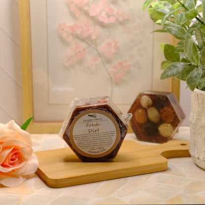 A hexagonal packaged soap labeled 'Fresh Dirt' sits on a wooden board on a tabletop, styled with a peach rose, a potted green plant, and a framed floral print in the background.