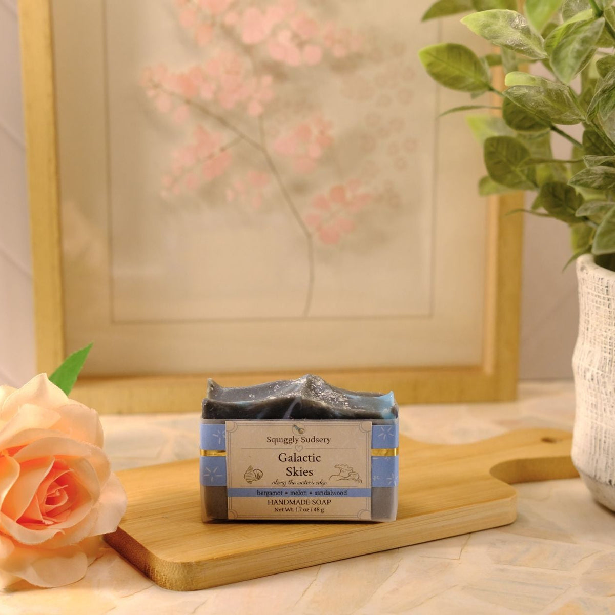 A bar of soap labeled 'Squiggly Sudsery Galactic Skies' sits on a wooden cutting board, styled with a peach rose, a potted plant, and a framed print in the background.