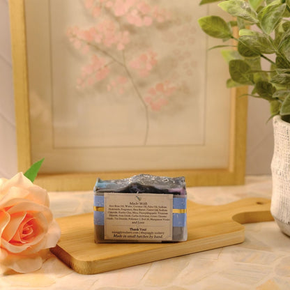 A packaged bar of soap sits on a wooden cutting board on a tabletop, styled with a peach rose, a potted green plant, and a framed floral print in the background.