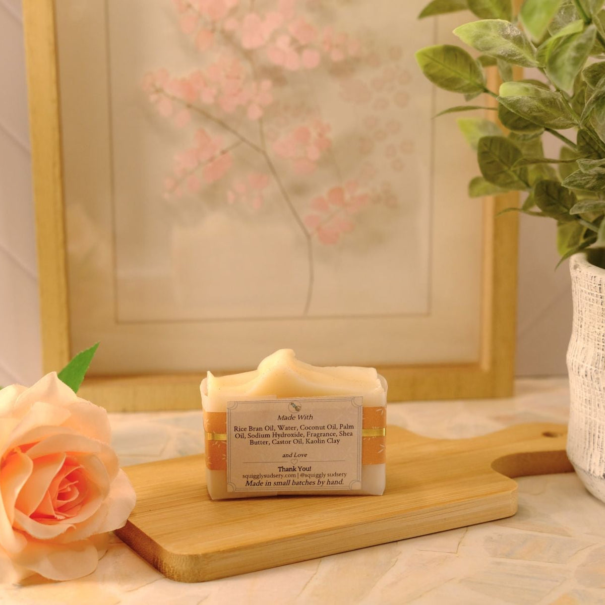 A bar of handmade soap sits on a wooden cutting board, styled with a peach rose, a potted green plant, and a framed floral print in a bright, clean setting.