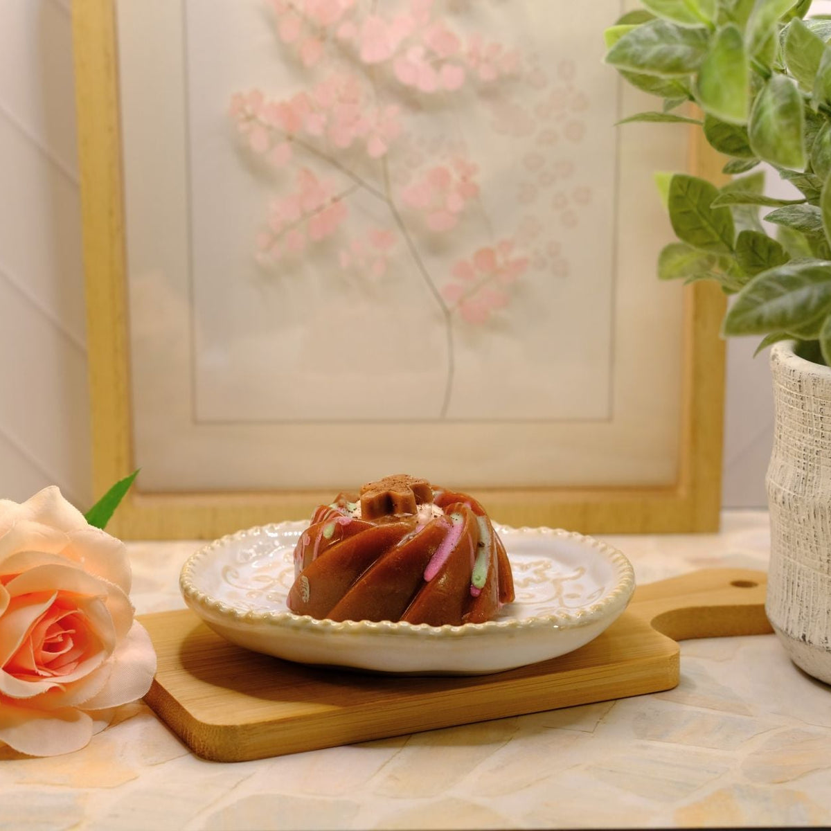 A small brown bundt cake soap on a white plate with a gold rim, set on a wooden cutting board on a patterned tablecloth. A peach rose, a potted green plant, and a framed floral print are arranged around it.