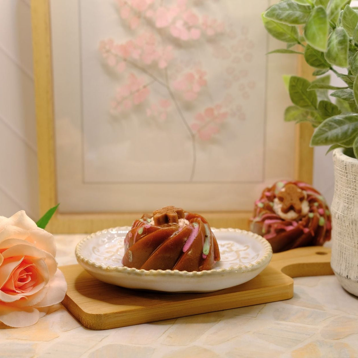 A small brown bundt cake soap sits on a white plate on a wooden board, styled on a tabletop with a peach rose, a potted plant, and a framed floral print in the background.