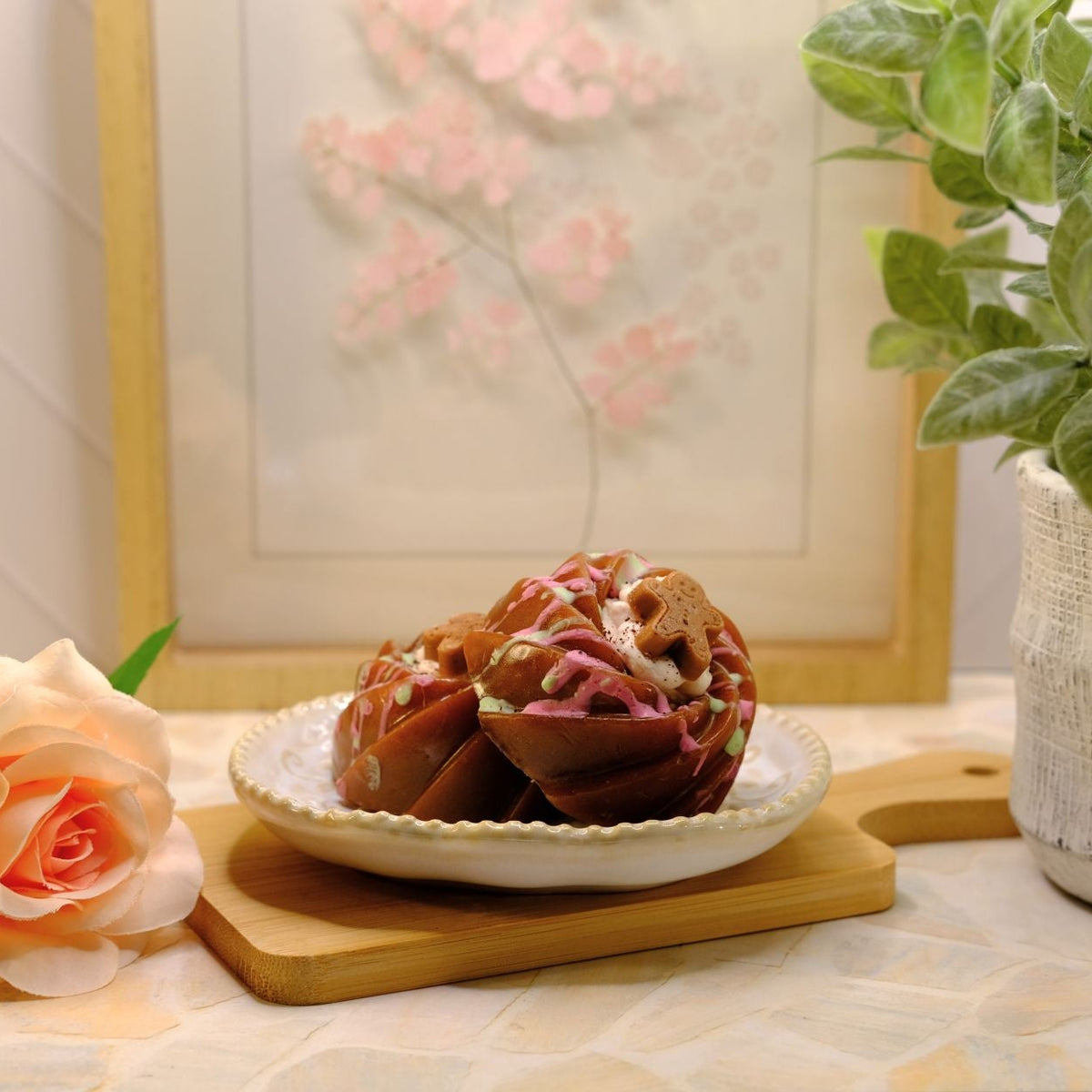 A bundt cake soap, placed on a small white plate on a wooden board. The setting includes a peach rose, a potted green plant, and a framed floral print in the background.