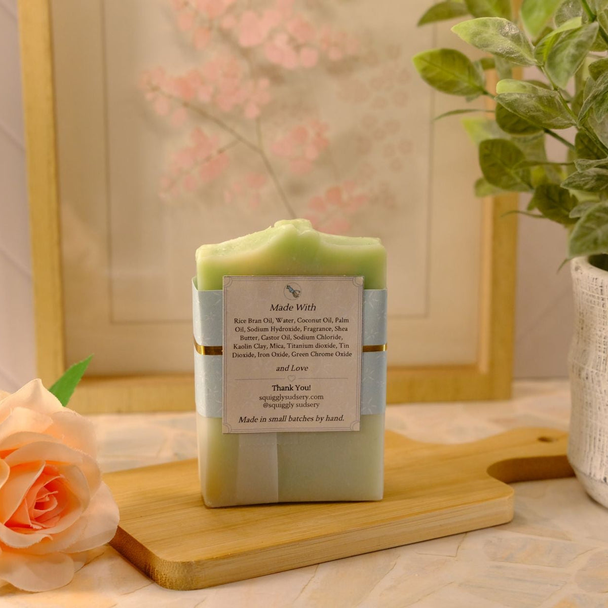 A handmade soap bar wrapped in a light blue paper sleeve with a white label, displayed on a wooden cutting board. The setting is a styled tabletop with a peach rose, a potted green plant, and a framed picture in the background.