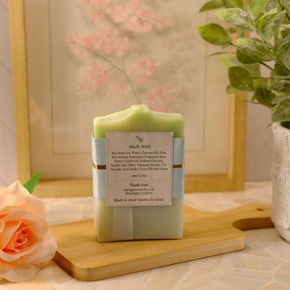 A handmade soap bar wrapped in a light blue paper sleeve with a white label, displayed on a wooden cutting board. The setting is a styled tabletop with a peach rose, a potted green plant, and a framed picture in the background.