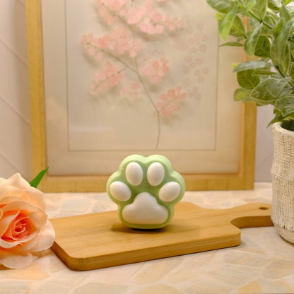 A small paw-shaped soap in mint green and white sits on a wooden cutting board on a tabletop, styled with a peach rose, a potted plant, and a framed floral print in the background.