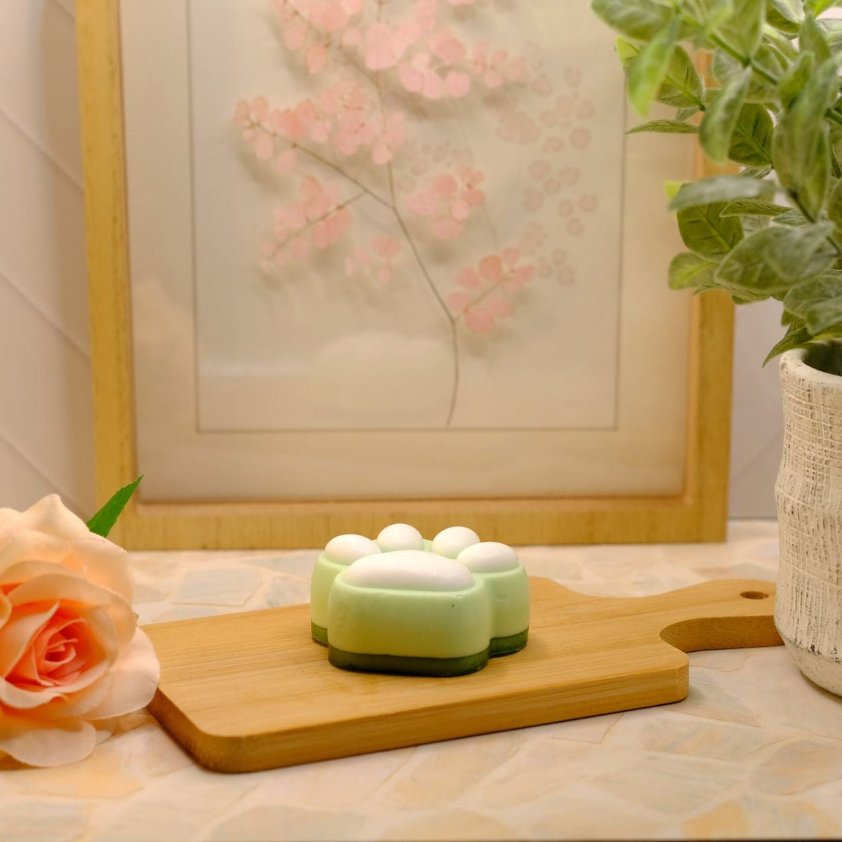 A small green and white paw-shaped soap sits on a wooden cutting board on a patterned surface, styled with a peach rose, a framed floral print, and a potted plant.