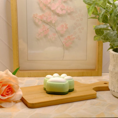 A small green and white paw-shaped soap sits on a wooden cutting board on a patterned surface, styled with a peach rose, a framed floral print, and a potted plant.