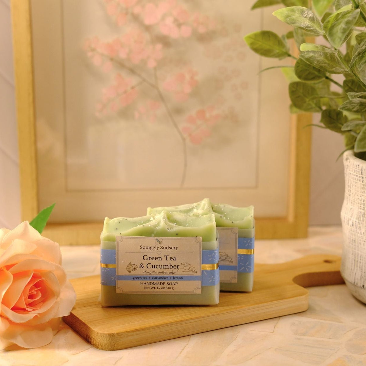 Two bars of soap labeled 'Green Tea & Cucumber' by Squiggly Sudsery, displayed on a wooden cutting board. The scene is styled with a peach rose on the left and a potted green plant on the right, set against a warm, soft-focus background.