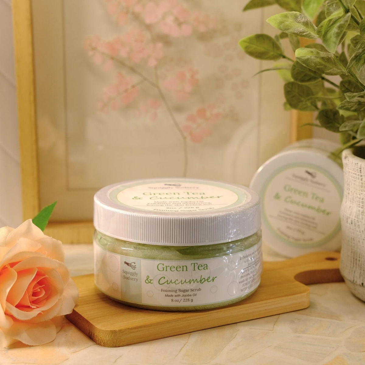 A jar of Green Tea & Cucumber sugar scrub sits on a wooden cutting board on a countertop, styled with a peach rose, a potted plant, and a second jar in the background.
