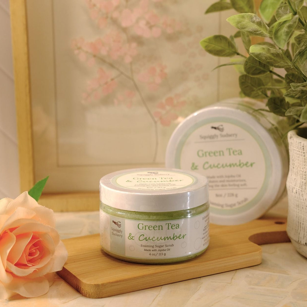 A jar of Green Tea & Cucumber sugar scrub sits on a wooden board next to a peach rose, with a second round jar and a potted plant in the background.