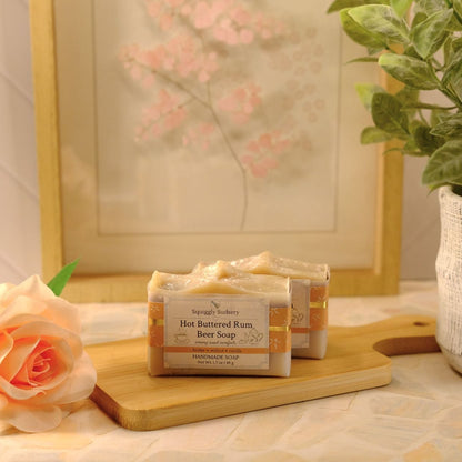 A bar of Squiggly Sudsery Hot Buttered Rum Bar Soap sits on a wooden cutting board next to a peach rose, with a potted green plant and a framed print in the background.