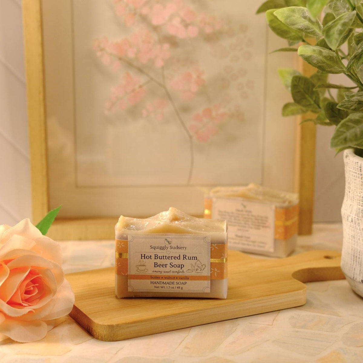 A bar of Squiggly Sudsery Hot Buttered Rum Beer Soap resting on a wooden cutting board, styled with a peach rose, a potted green plant, and a framed print in the background.