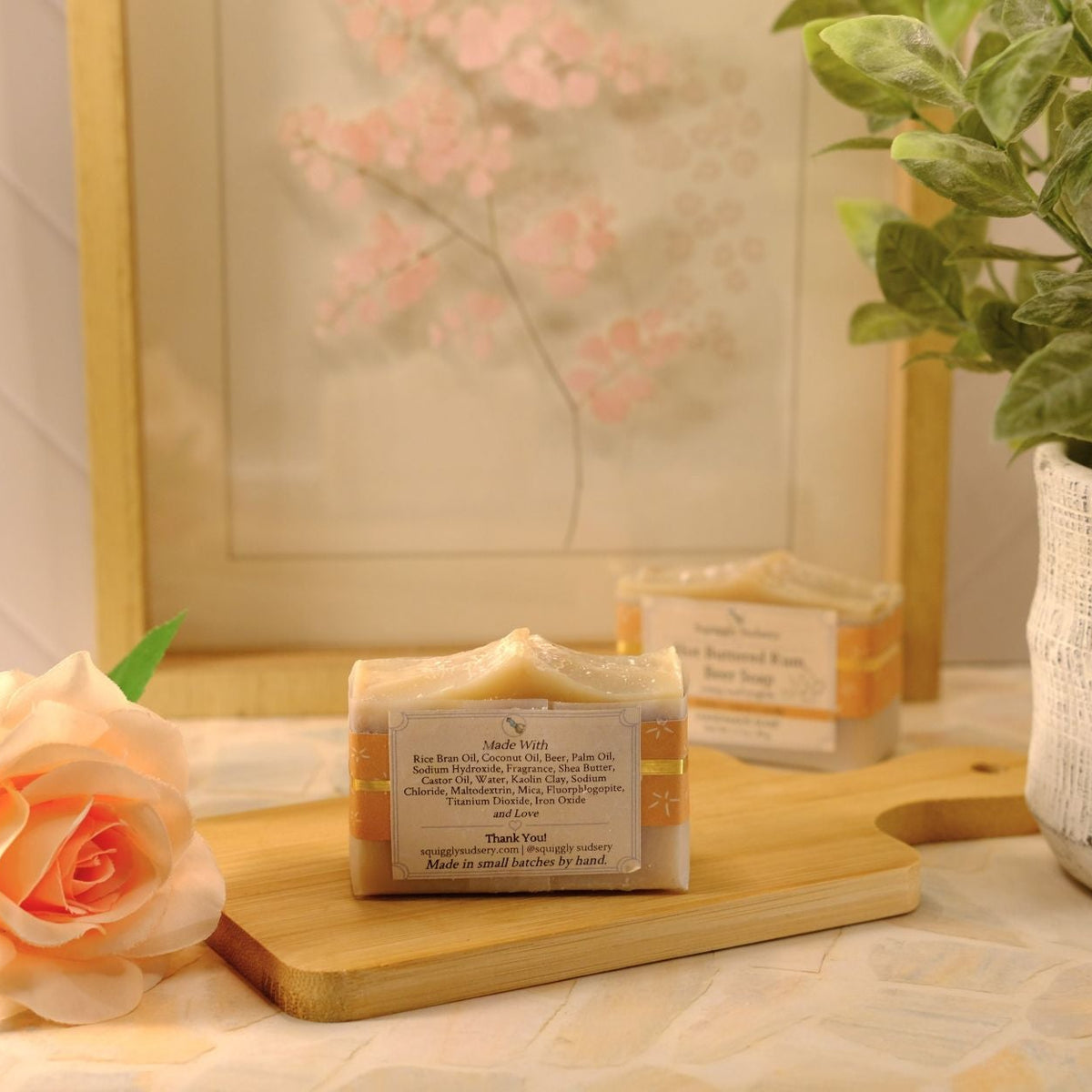 A handmade soap bar with a cream and gold label sits on a wooden cutting board, styled with a peach rose, a potted green plant, and a framed floral print in the background.
