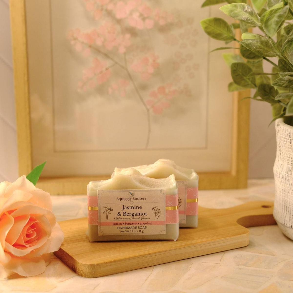 Two wrapped bars of soap labeled Jasmine & Bergamot sit on a wooden cutting board on a tiled countertop, styled with a peach rose, a potted green plant, and a framed floral print in the background.