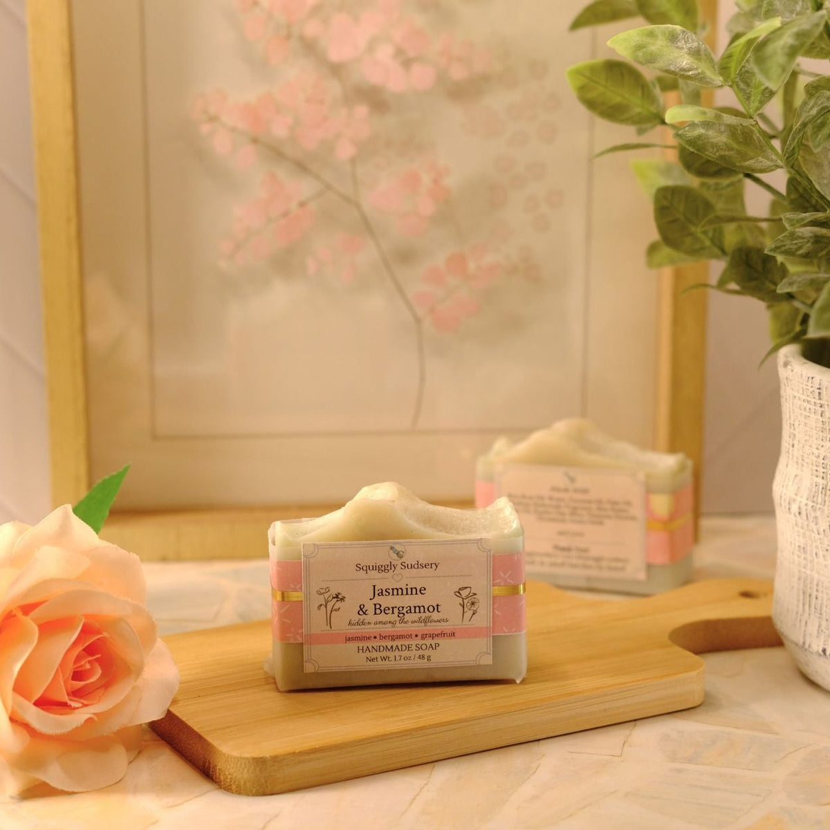 A bar of soap labeled 'Jasmine & Bergamot' by Squiggly Sudsery sits on a wooden cutting board on a tiled countertop, styled with a peach rose, a potted plant, and a framed print in the background.