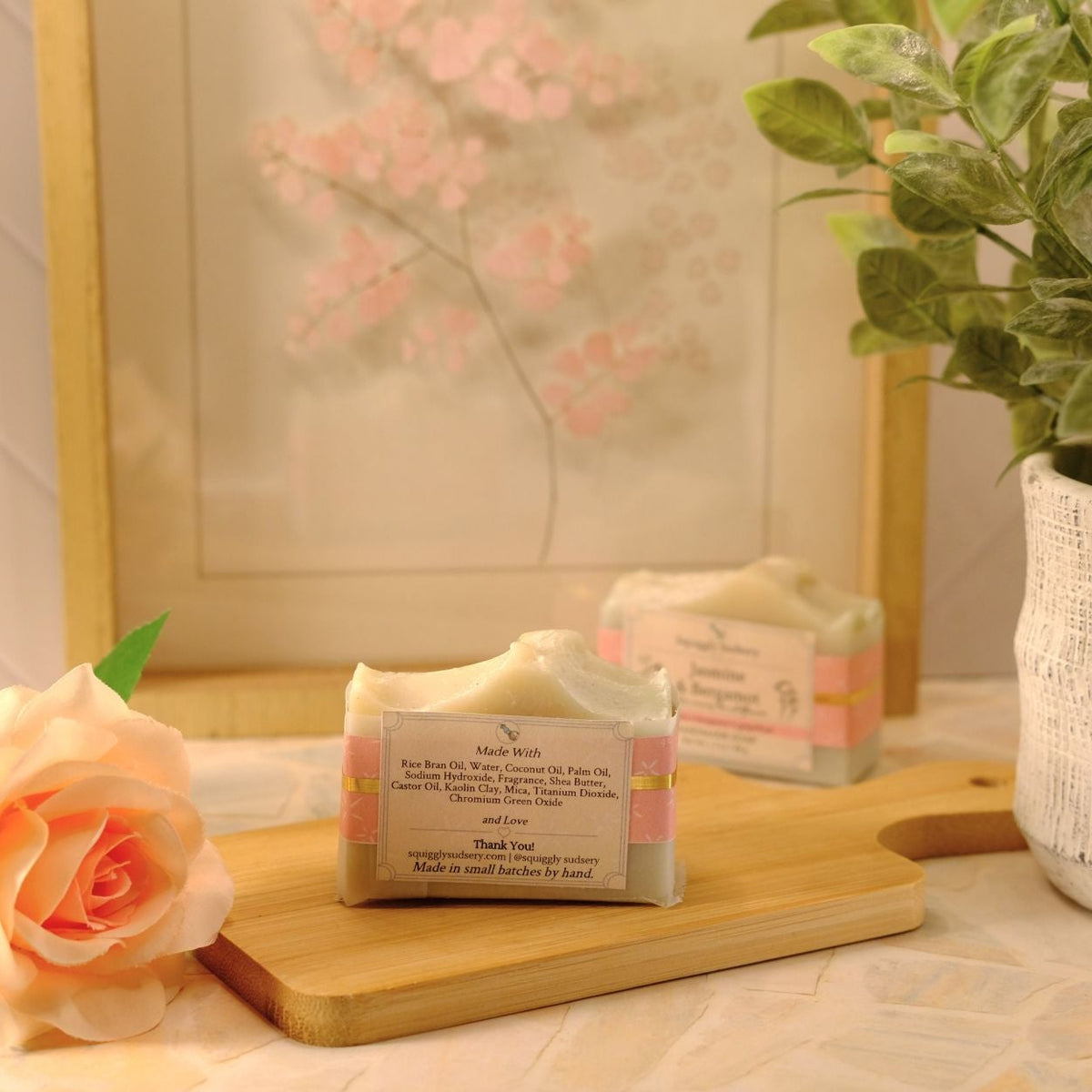 A bar of soap wrapped in a kraft paper sleeve sits on a wooden cutting board on a table, styled with a peach rose, a potted plant, and a framed print in the background.
