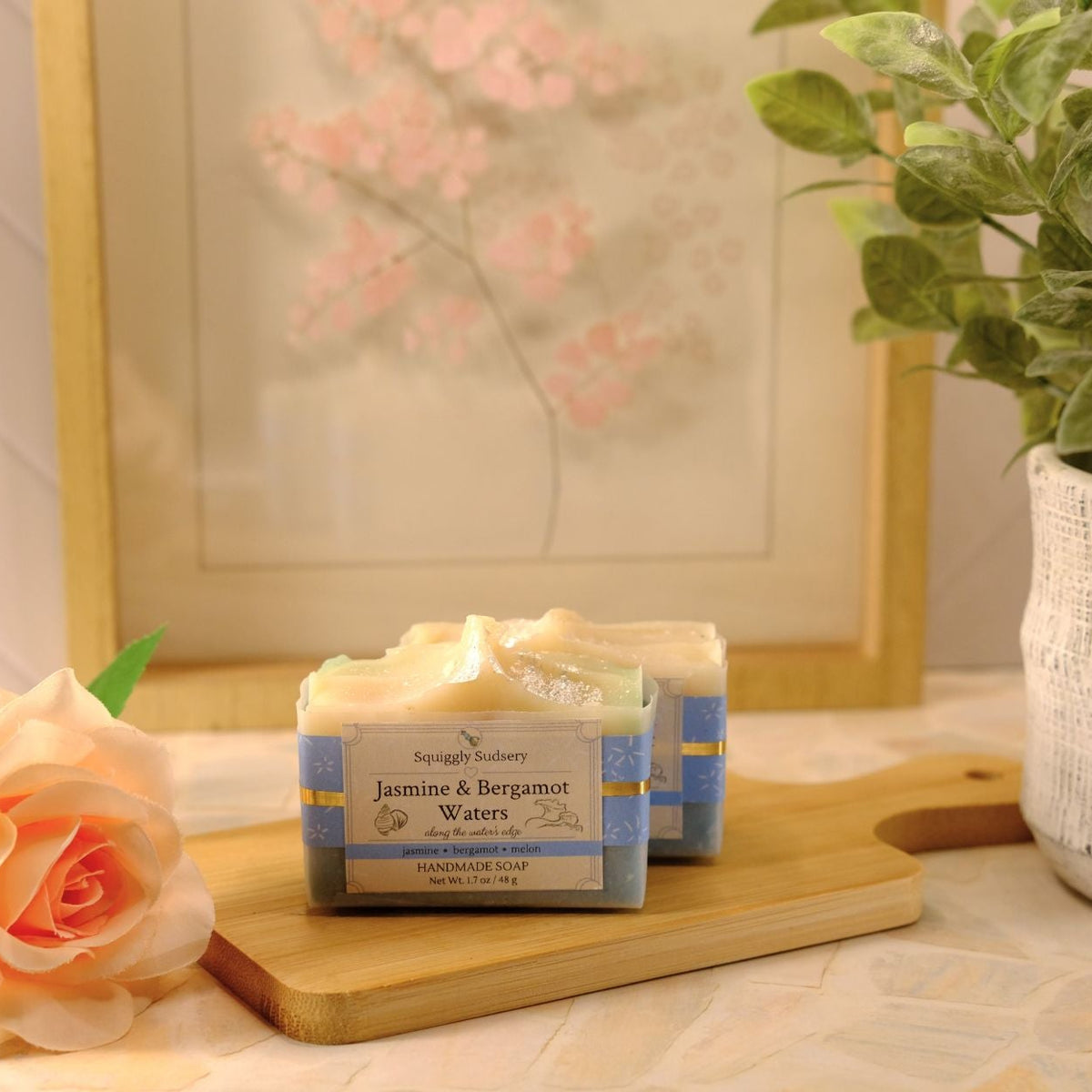Two bars of handmade soap labeled 'Jasmine & Bergamot Water' by Squiggly Sudsery, styled on a wooden cutting board with a peach rose and a potted plant in a warm setting.