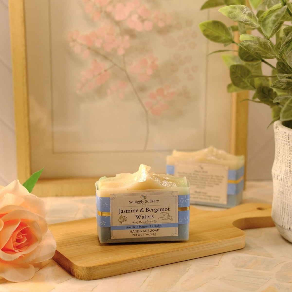 A rectangular bar of soap labeled 'Jamine & Bergamot Waters' sits on a wooden board on a tabletop, styled with a peach rose, a potted green plant, and a framed floral print in the background.