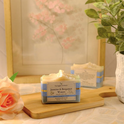 A rectangular bar of soap labeled 'Jamine & Bergamot Waters' sits on a wooden board on a tabletop, styled with a peach rose, a potted green plant, and a framed floral print in the background.