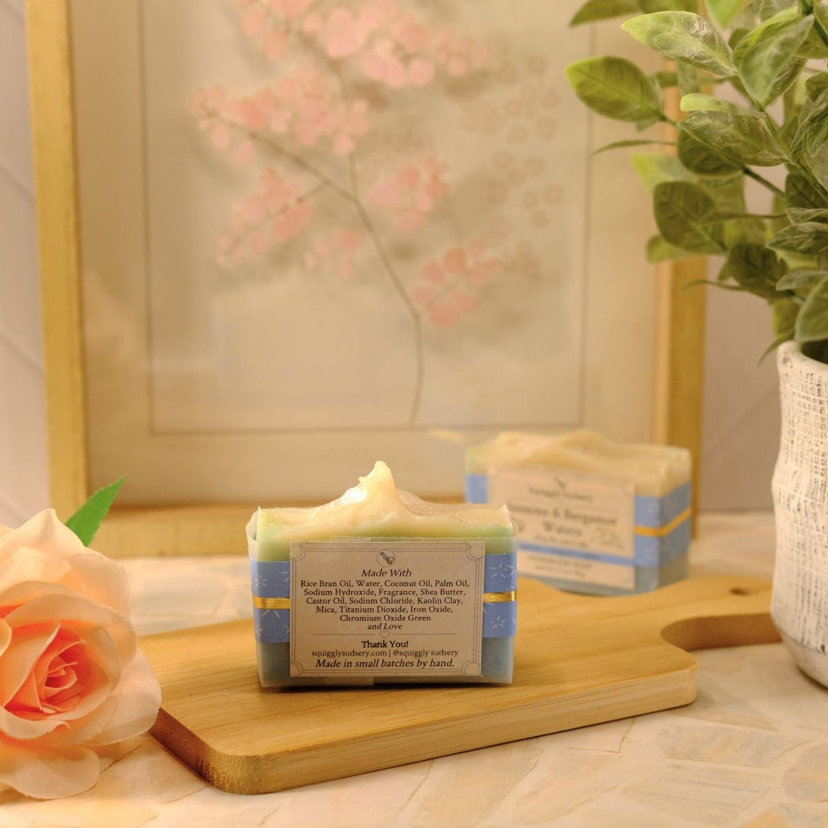 A handmade soap bar resting on a wooden cutting board, styled with a peach rose, a potted plant, and a framed print in a warm, soft-focus setting.