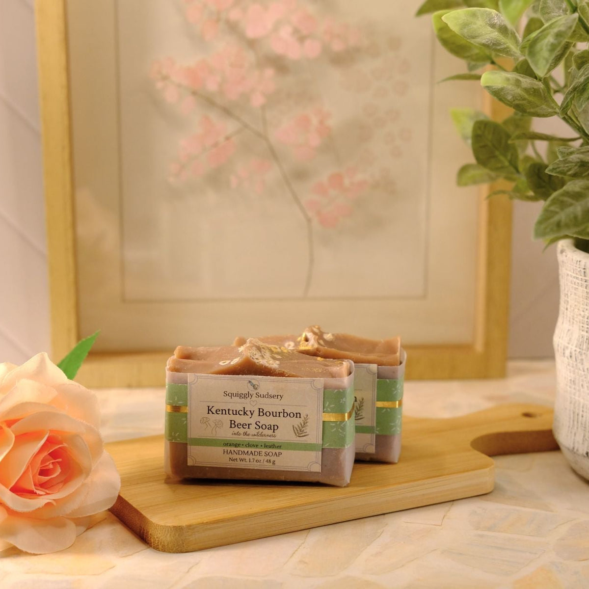 Two bars of Kentucky Bourbon Beer Soap displayed on a wooden cutting board, styled with a peach rose, a potted green plant, and a framed floral print in the background.