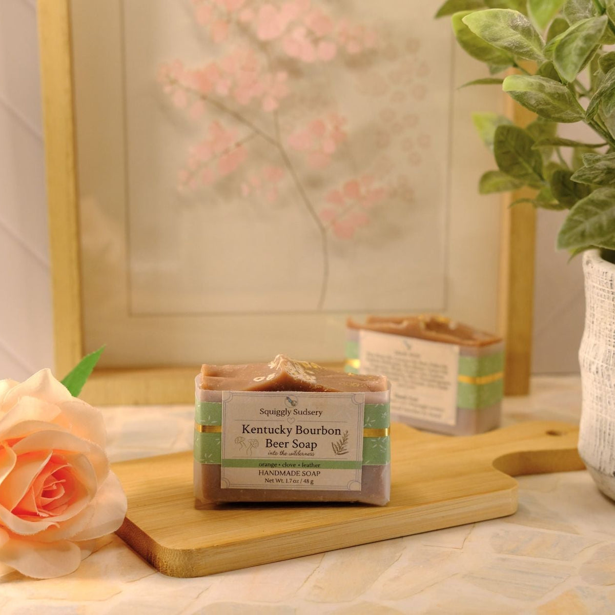 A bar of Kentucky Bourbon Beer Soap sits on a wooden cutting board on a tiled countertop, styled with a peach rose, a potted green plant, and a framed floral print in the background.