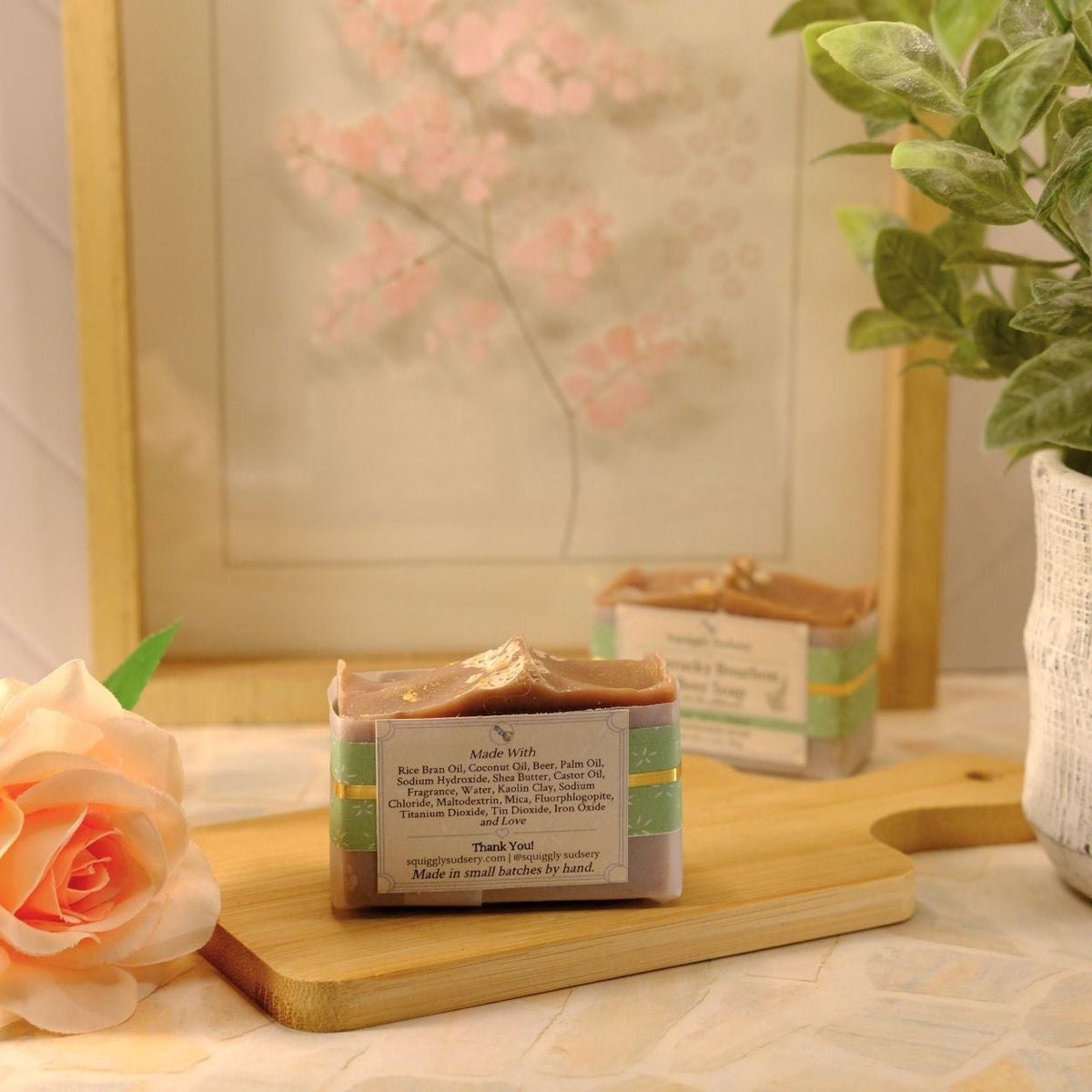 A bar of soap sits on a wooden cutting board on a tiled countertop, styled with a peach rose, a potted green plant, and a framed floral print in the background.