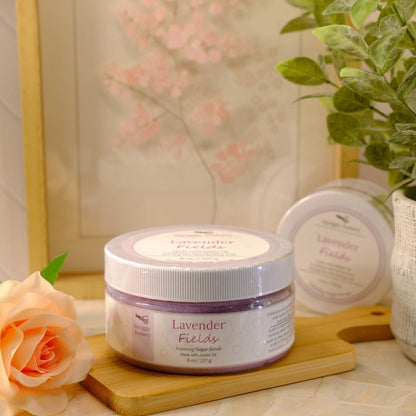 Lavender Fields sugar scrub in a clear jar with a white lid, displayed on a wooden board next to a peach rose and a potted green plant.