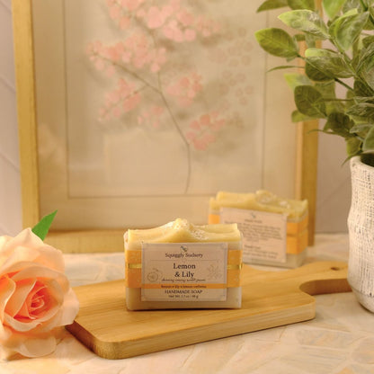 A bar of soap labeled 'Lemon & Lily' by Squiggly Sudsery, sits on a wooden cutting board, styled with a peach rose, a potted plant, and a framed print in the background.