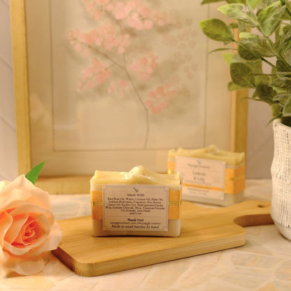 A handmade soap bar wrapped in kraft paper packaging sits on a wooden cutting board, styled with a peach rose, a potted plant, and a framed print in a warm, soft-focus setting.