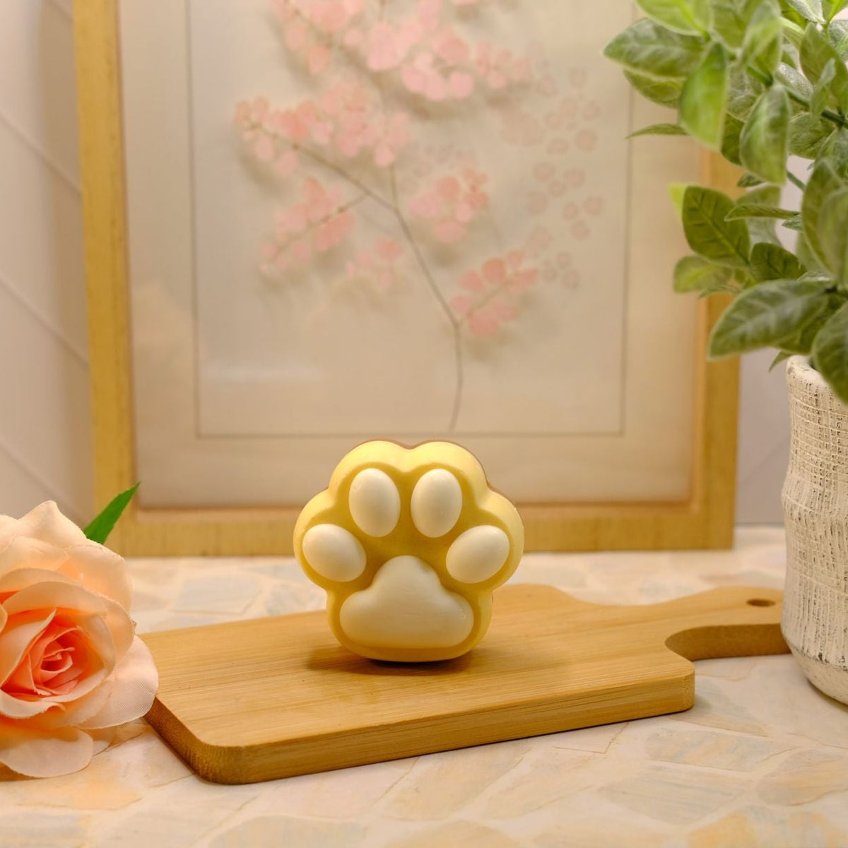 A yellow and white cat paw-shaped soap sits on a wooden cutting board, styled with a peach rose, a potted green plant, and a framed picture in the background.