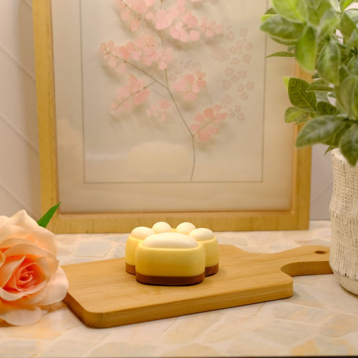 Small paw shaped soap laying down on a wooden cutting board, styled with a peach rose, a potted green plant, and a framed floral print in the background.