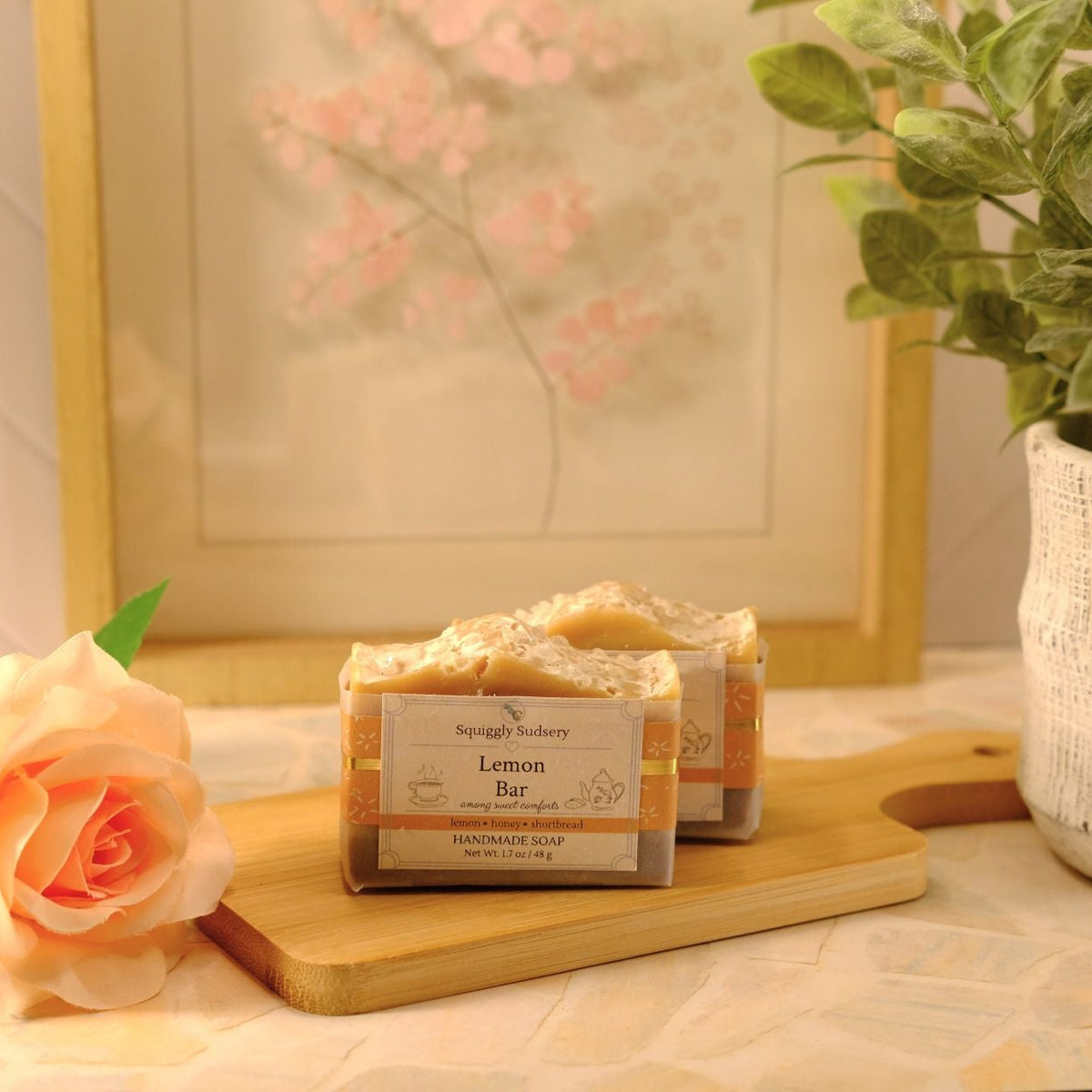 Two wrapped soap bars labeled 'Lemon Bar' by Squiggly Sudsery, displayed on a wooden cutting board with a peach rose and a potted plant in a softly lit indoor setting.