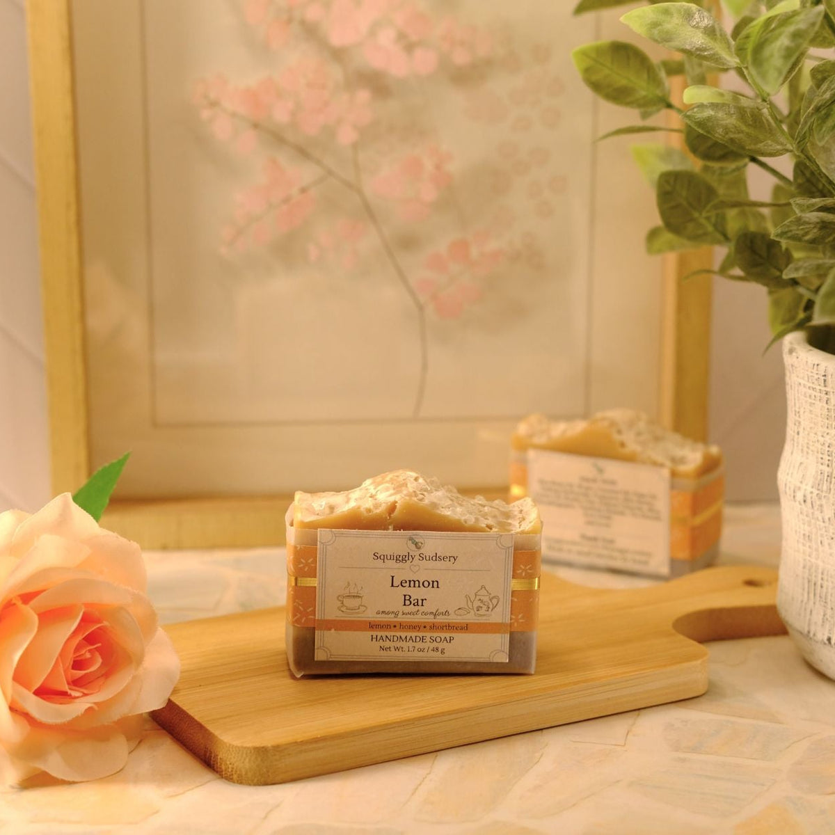 A rectangular bar of soap labeled 'Lemon Bar' sits on a wooden cutting board on a table, styled with a peach rose, a potted plant, and a framed print in a warm, soft-focus setting.