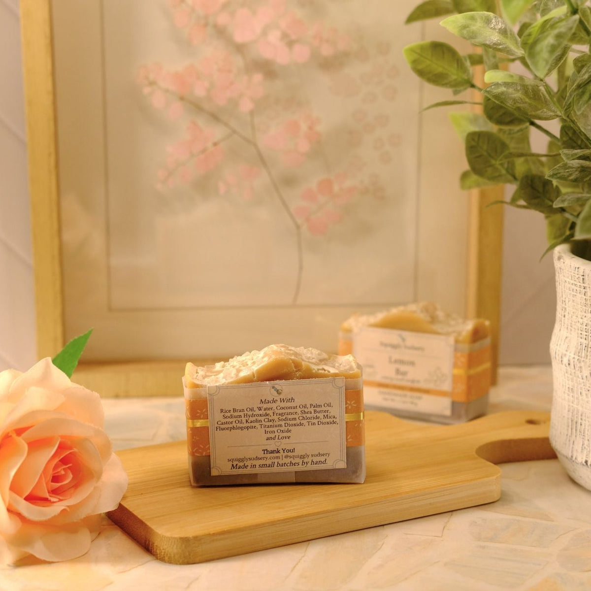 Two rectangular bar soaps with cream-colored tops and orange-brown striped sides, displayed on a wooden cutting board with a peach rose and a potted plant in a softly lit indoor setting.
