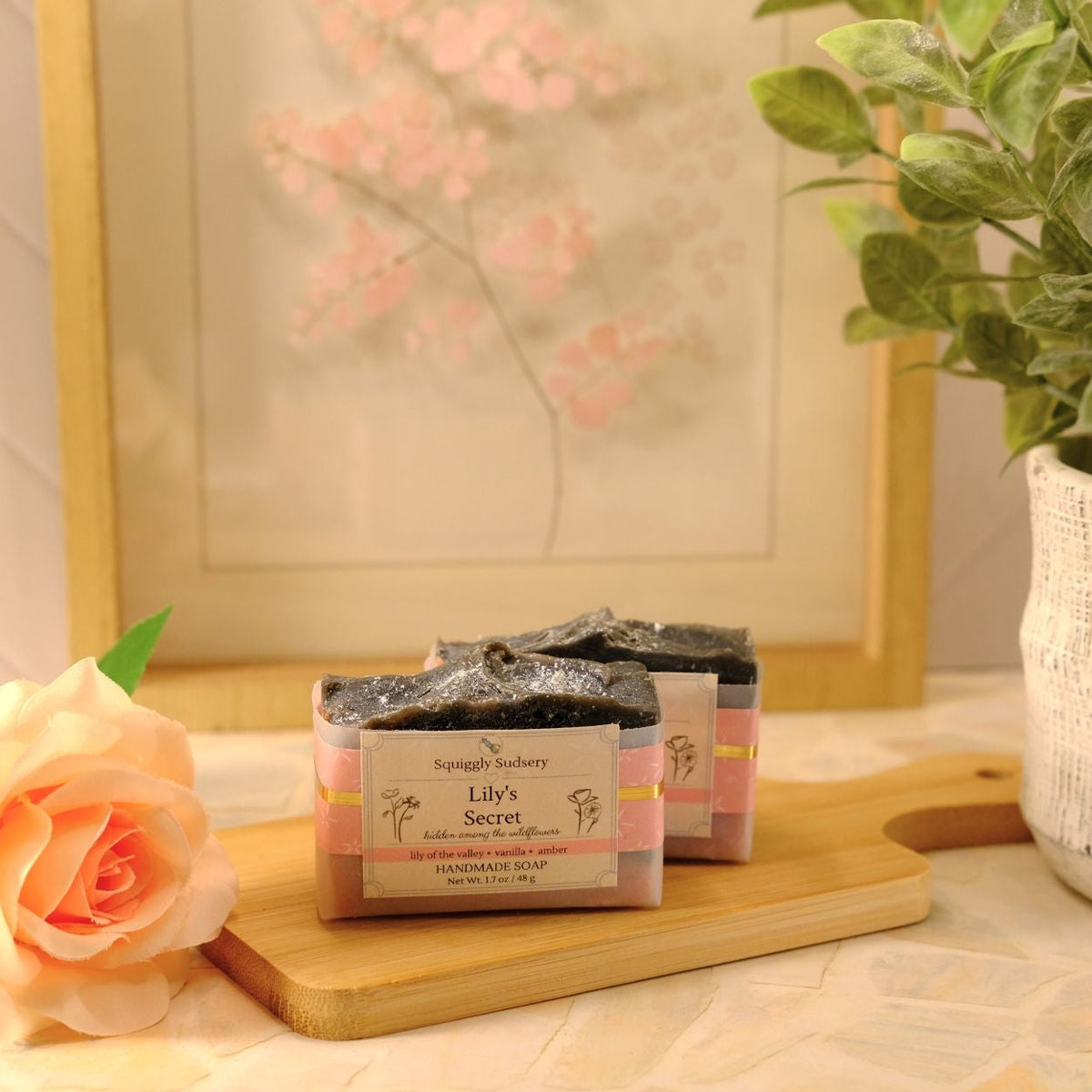 Two packaged soap bars labeled 'Lily's Secret' sit on a wooden cutting board on a tiled countertop, styled with a peach rose, a potted green plant, and a framed floral print in the background.