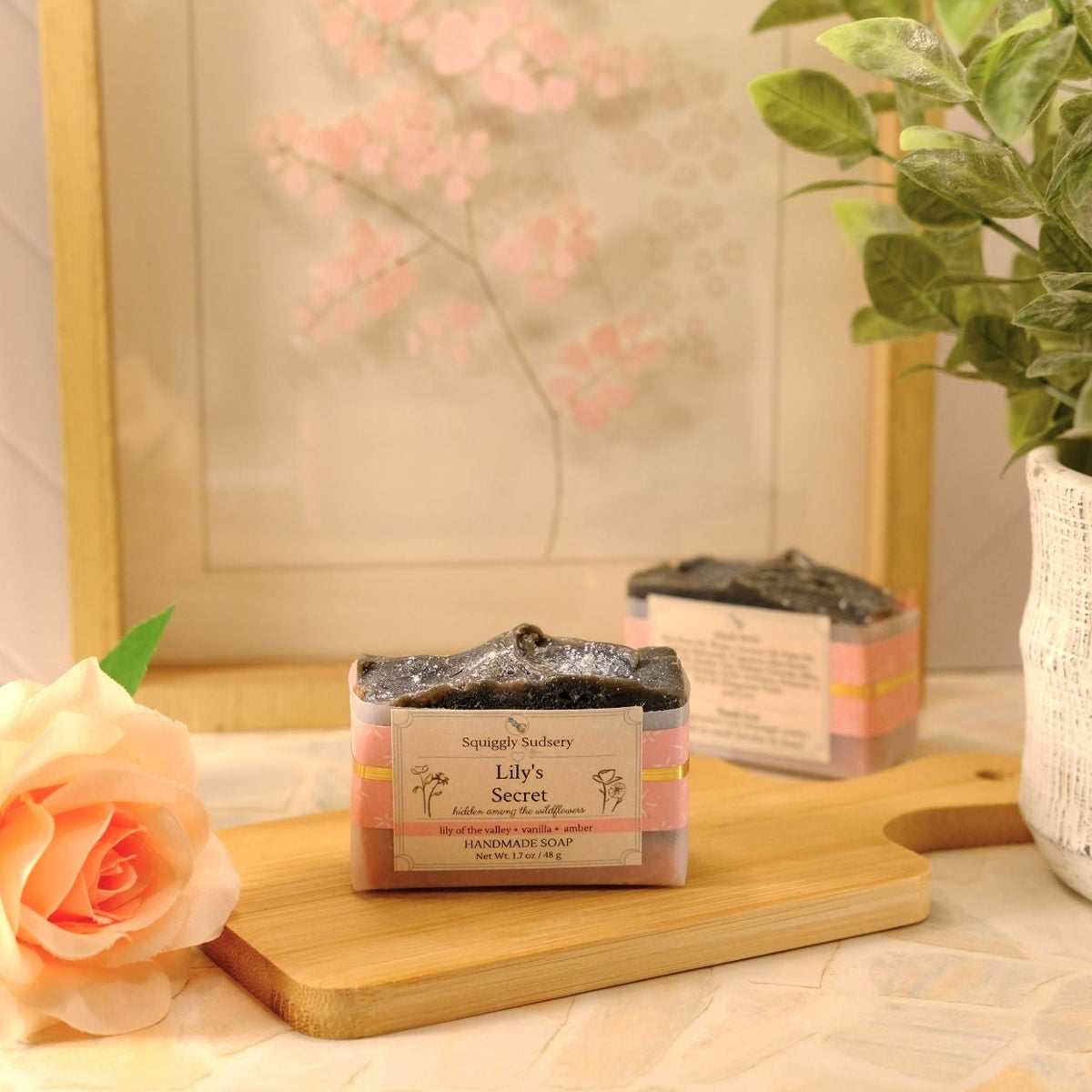 A styled product shot of two artisan soap bars on a wooden cutting board, surrounded by decorative props including a peach rose, a potted green plant, and a framed floral print.