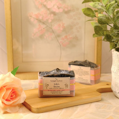 A styled product shot of two artisan soap bars on a wooden cutting board, surrounded by decorative props including a peach rose, a potted green plant, and a framed floral print.
