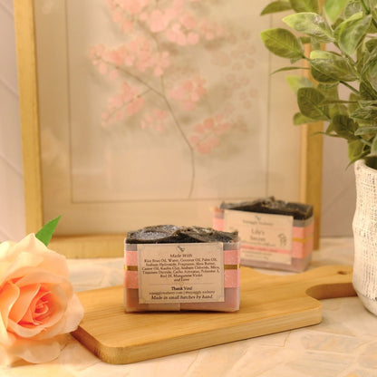 A small rectangular soap bar with a pink and white label sits on a wooden cutting board, with a second soap bar behind it and a pink rose in the foreground. A potted green plant and a framed floral print are visible in the softly lit background.