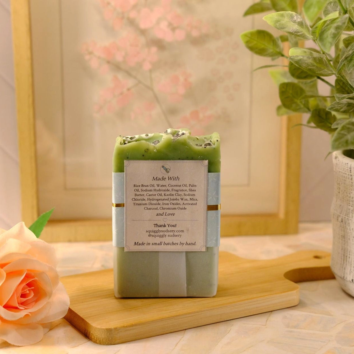 A handmade soap bar sits on a wooden cutting board. The soap has a sage green base with a lime green top layer decorated with small black dots.