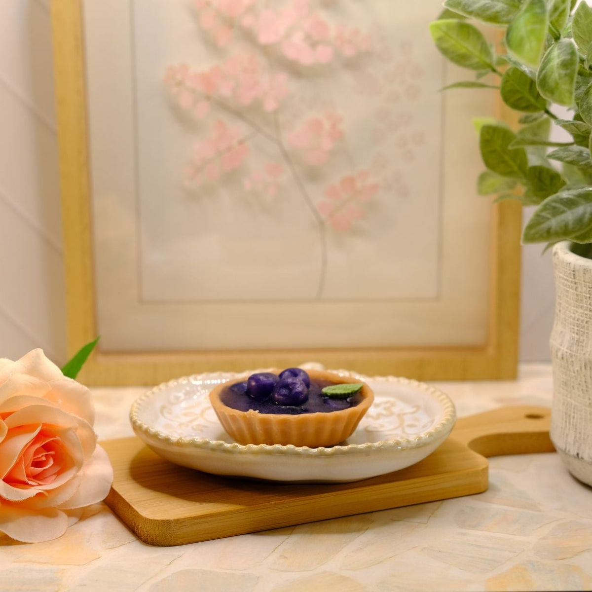 A small soap tart with dark purple berries on a white plate, styled on a wooden board with a peach rose and a potted green plant in a softly lit indoor setting.